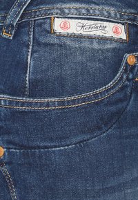 Mörkblå denimjeans med en vit etikett med röda detaljer som läser "Herrlicher." Inkluderar distinkt sömnad och en knappdetalj.