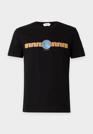 Mężczyzna ubrany w czarną koszulkę Versace z logo Medusa, szare szerokie spodnie w paski, czarne buty i okulary przeciwsłoneczne na białym tle.
