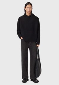 Czarna, oversize'owa bluza z kapturem o fakturowanym wykończeniu, zestawiona z czarnymi spodniami typu wide-leg. Modelka trzyma ciemną skórzaną torbę i nosi czarne buty.