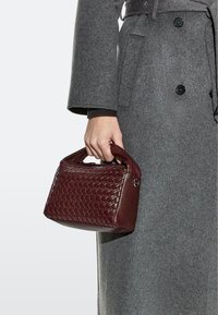 Hand hält eine kleine dunkelrote geflochtene Lederhandtasche, trägt einen langen grauen Mantel mit schwarzen Knöpfen.