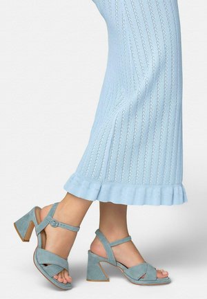 Piernas con pantalones de punto azul claro y sandalias de tacón bloque de ante azul claro a juego con tiras en el tobillo sobre un fondo blanco.