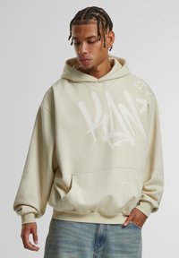 Sudadera beige hecha de tejido suave, con corte holgado, grandes gráficos en estilo graffiti en blanco, bolsillo frontal y puños acanalados.
