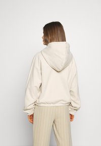 Femme aux cheveux bruns mi-longs portant un sweat à capuche beige et un pantalon beige à fines rayures, vue de dos sur un fond blanc uni.