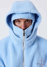 Personne portant une veste à capuche pelucheuse bleu clair, zippée jusqu'au nez, ne montrant que ses yeux et une partie de son visage.