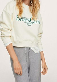 Felpa crema con stampa verde "SPORTS CLUB", con polsini e orlo a costine. Abbinata a pantaloni jogger grigi, con una vestibilità comoda.