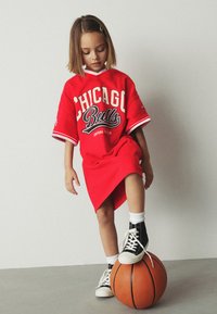 Maglia sportiva rossa oversize con logo "Chicago Bulls", abbinata a scarpe da ginnastica nere alte. L'ambientazione include un pallone da basket.