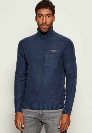 Maglione a collo alto blu navy con polsini e orlo a costine, dotato di tasca a toppa e logo ricamato in oro sul petto. Tessuto a maglia liscia.