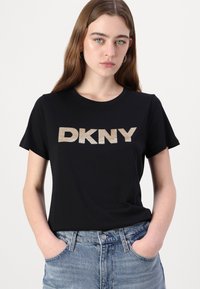 T-shirt noir en coton à manches courtes avec un logo "DKNY" doré métallisé sur l'avant. Assorti à un jean taille haute bleu clair.