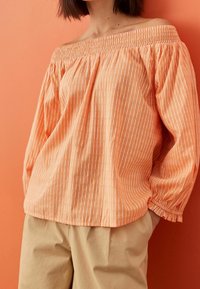Orange off-the-shoulder blouse with white vertical stripes, elasticized neckline, and gathered cuffs, paired with beige loose-fitting pants.