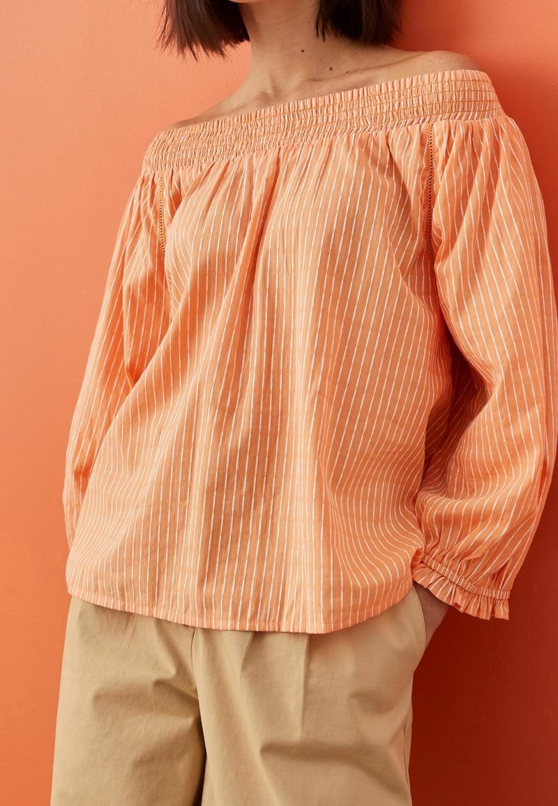 Orange off-the-shoulder blouse with white vertical stripes, elasticized neckline, and gathered cuffs, paired with beige loose-fitting pants.