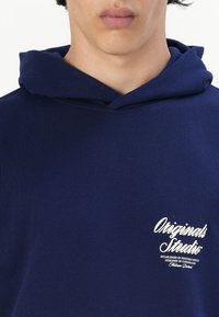 Donkerblauwe hoodie van gladde stof, met een voorvak en het "Originals Studio" logo in het wit op de linkerkant van de borst.