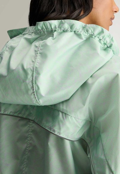 Kvinde med mørkt hår iført en lysegrøn regnjakke med stor hætte og ventileret rygklap, vendt mod højre.