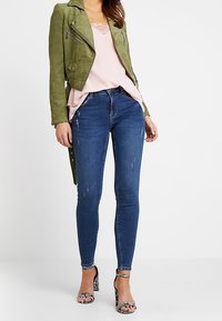 Chaqueta de motorista de ante verde, top de tirantes rosa claro con encaje, vaqueros ajustados azul oscuro y sandalias de tacón con patrón en blanco y negro.