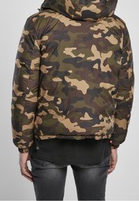 Padded camouflage jacket in green, brown, and beige tones. Features a hood, elastic cuffs, and a cinch detail at the hem.