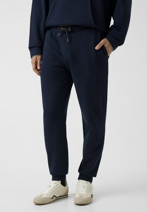 Uomo che indossa pantaloni da jogging blu navy con tasche laterali e scarpe da ginnastica bianche, in piedi davanti a uno sfondo chiaro e uniforme.