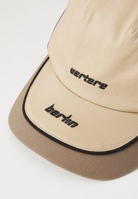 Casquette beige avec une visière brun foncé. Présente "vertebré" et "berlin" en broderie noire. Tissu texturé avec des détails de couture contrastés.