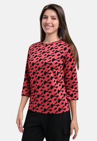 Red top with a black abstract pattern, round neck, and three-quarter sleeves. Made from a smooth fabric, paired with black pants.