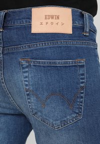 Niebieskie dżinsy mają brązową skórzaną metkę z napisem „EDWIN” oraz japońskim tekstem. Tylny kieszeń ma charakterystyczny krzywy wzór przeszycia.
