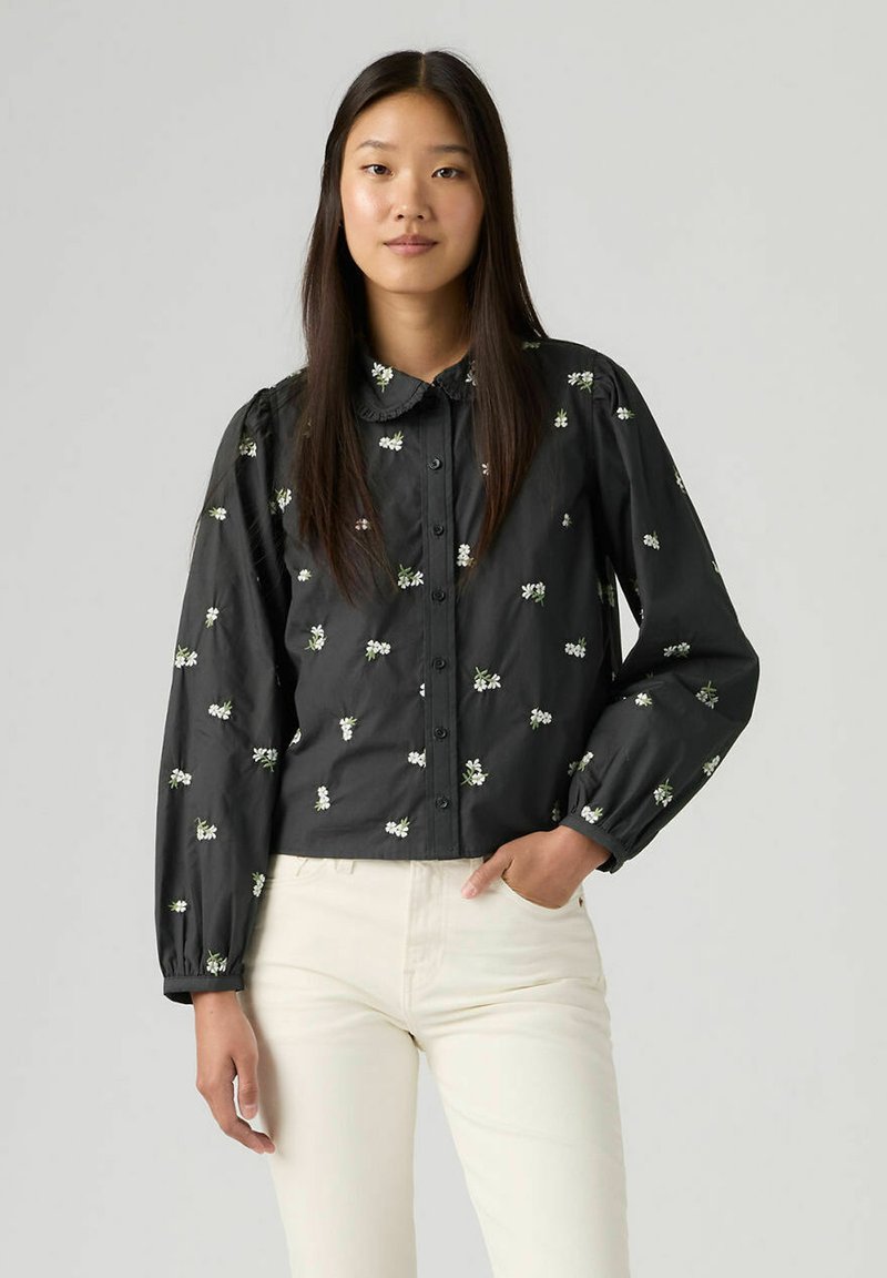 Chemise noire boutonnée avec col, ornée de broderies florales blanches, manches longues bouffantes et coupe légèrement courte. Portée avec un pantalon couleur crème.