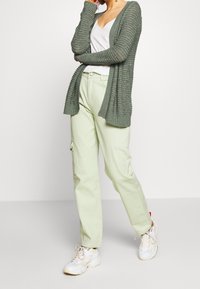 Osoba v zeleném strukturovaném kardiganu, bílém topu, světle zelených cargo kalhotách a bílých teniskách, stojící na pozadí s plain bílou.