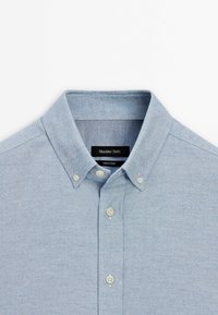 Chemise bleu clair à boutons avec un col pointu, trois boutons blancs et une étiquette noire à l’intérieur du col indiquant Massimo Dutti.