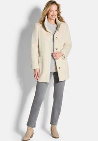 Crème-kleurige, gestructureerde jas met een hoge kraag, voorzien van knoopsluitingen en zijzakken, gecombineerd met grijze broek en beige enkellaarzen.