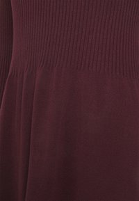 Pull en tricot bourgogne avec un corps côtelé et un bas évasé lisse. Le tissu a une finition douce et texturée. Les détails sont subtils et uniformes.