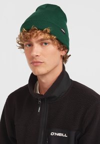 Groene geribbelde gebreide beanie van zachte stof, gedragen met een zwart fleece jack met rits en logo op de borst.