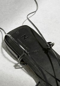 Sandale en cuir noir avec des lanières fines et un logo embossé sur la semelle, posée sur une surface claire et texturée.