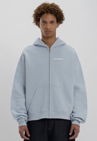 Felpa zip-up azzurro chiaro con tasca frontale, vestibilità morbida, polsini a costine e cappuccio. Presenta un branding discreto sul petto in bianco.
