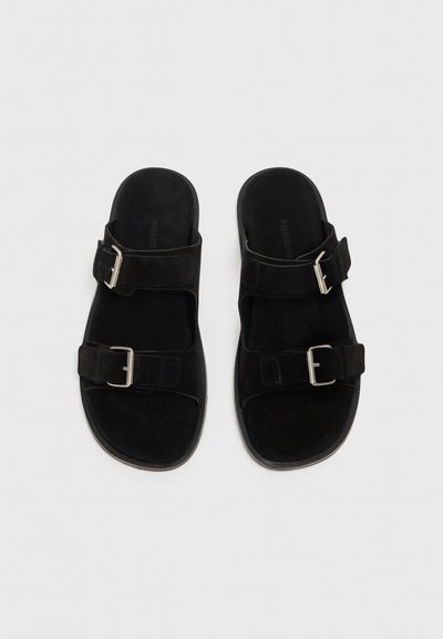Sandali in suede nero con due cinturini regolabili, fibbie in argento e suola piatta in gomma. Presentano una texture liscia e un design minimale.