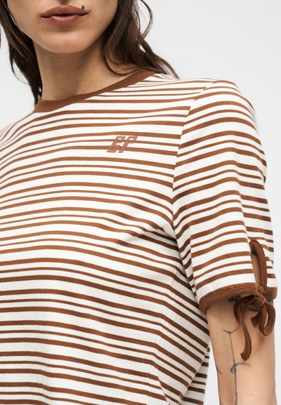 Γυναίκα που φοράει μια κοντομάνικη μπλούζα με λευκές και καφέ οριζόντιες ρίγες, με μικρό λογότυπο και λεπτομέρεια μανικοδέτη με φιόγκο, εμφανίζοντας μέρος ενός τατουάζ στο μπράτσο.