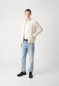 Ljusbeige bomberjacka med en slät yta, kombinerad med ljusblå jeans och svarta snörskor. Jackan har en liten logotypdetalj.
