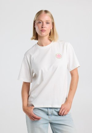 Staande vrouw met kort blond haar, die een wit T-shirt draagt met een klein roze en groen logo op de borst en lichtblauwe spijkerbroek.