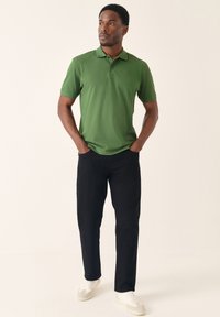 Zöld rövid ujjú galléros póló finom logóval; fekete nadrággal és fehér sportcipővel párosítva. Egyszerű kialakítás és szabás.