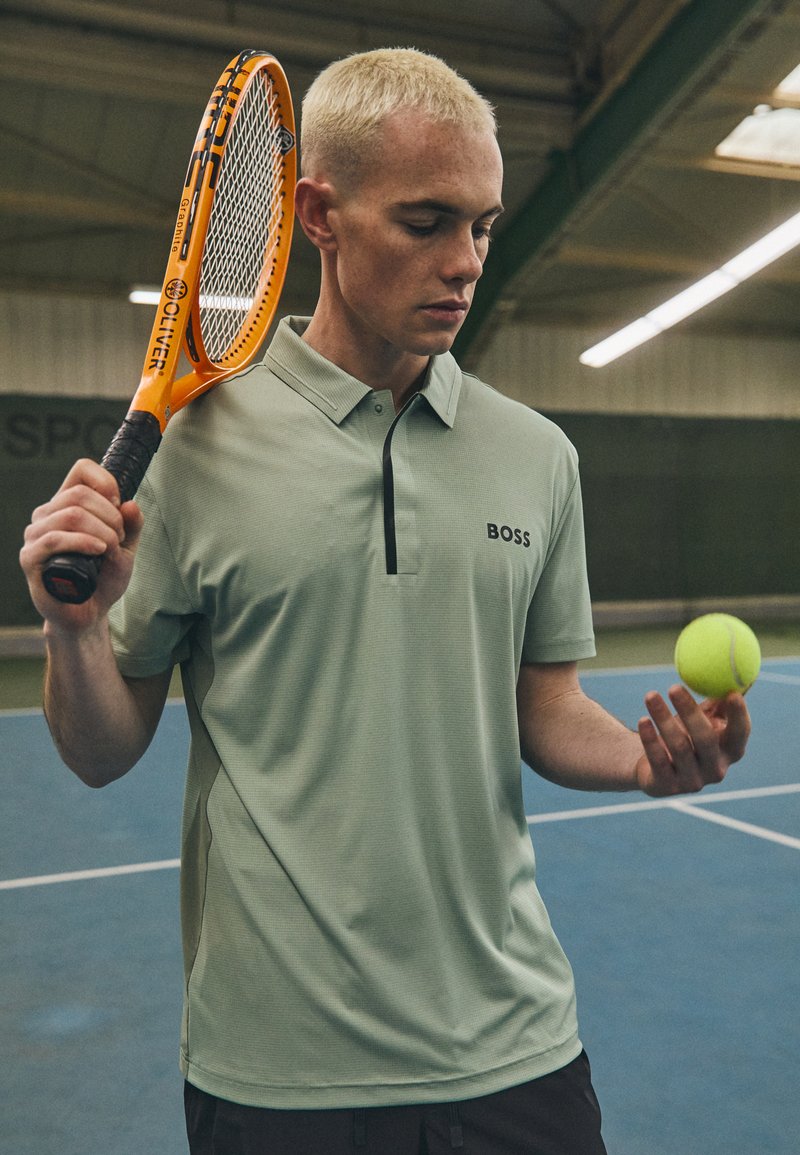 Vihreä, lyhythihainen polo-paita, jossa on napitettava kaulus ja musta yksityiskohta, jota henkilö pitää tennisracketin ja pallon kanssa kentällä.