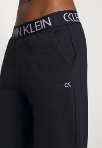 Zbliżenie na osobę ubraną w czarne dresy Calvin Klein z logo na pasie i małym logo CK na udzie.