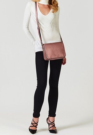 Kobieta w białej bluzce z długim rękawem, czarnych obcisłych spodniach, czarnych sandałkach na obcasie z paskami, nosząca na ramieniu różowo-złotą torebkę na pasku.