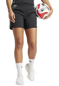 Zwarte sportshorts met een elastische tailleband, gecombineerd met witte sokken en lichte sneakers, terwijl er een gemusterde voetbal wordt vastgehouden.