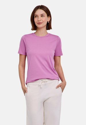 Jonge vrouw met kort bruin haar, gekleed in een roze T-shirt met korte mouwen en witte broek met trekkoord, handen in de zakken.