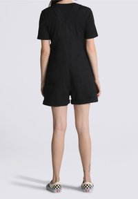 Person standing, wearing black short overalls, black t-shirt, and checkered slip-on sneakers, showing back view.