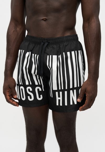Short de bain noir avec des rayures blanches verticales en code-barres et le nom de la marque écrit en lettres réparties à l'avant, porté par un mannequin masculin à la peau foncée, torse nu.