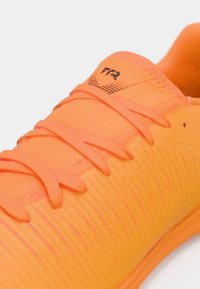 Jasnopomarańczowy but sportowy z fakturowaną cholewką, płaskimi sznurowadłami i subtelnym czarnym logo. Gładkie wykończenie i lekka konstrukcja.