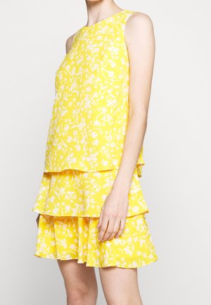 Ärmelloses gelbes Kleid mit mehrlagigem Rock und weißem Blumenmuster, getragen von einer stehenden Person vor neutralem Hintergrund.