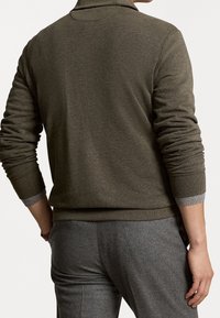 Homme portant un pull à manches longues vert olive et un pantalon gris, vu de dos avec les manches légèrement remontées.