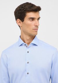 Chemise bleu clair à manches longues avec un col pointu, en tissu texturé et des détails de boutons décoratifs. Boutons marron avec un léger brillant.