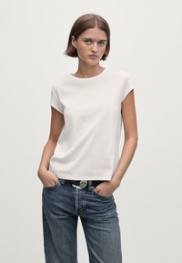 Donna con i capelli castani corti che indossa una maglietta bianca a maniche corte infilata in un paio di jeans blu larghi con una cintura nera, mani in tasca.
