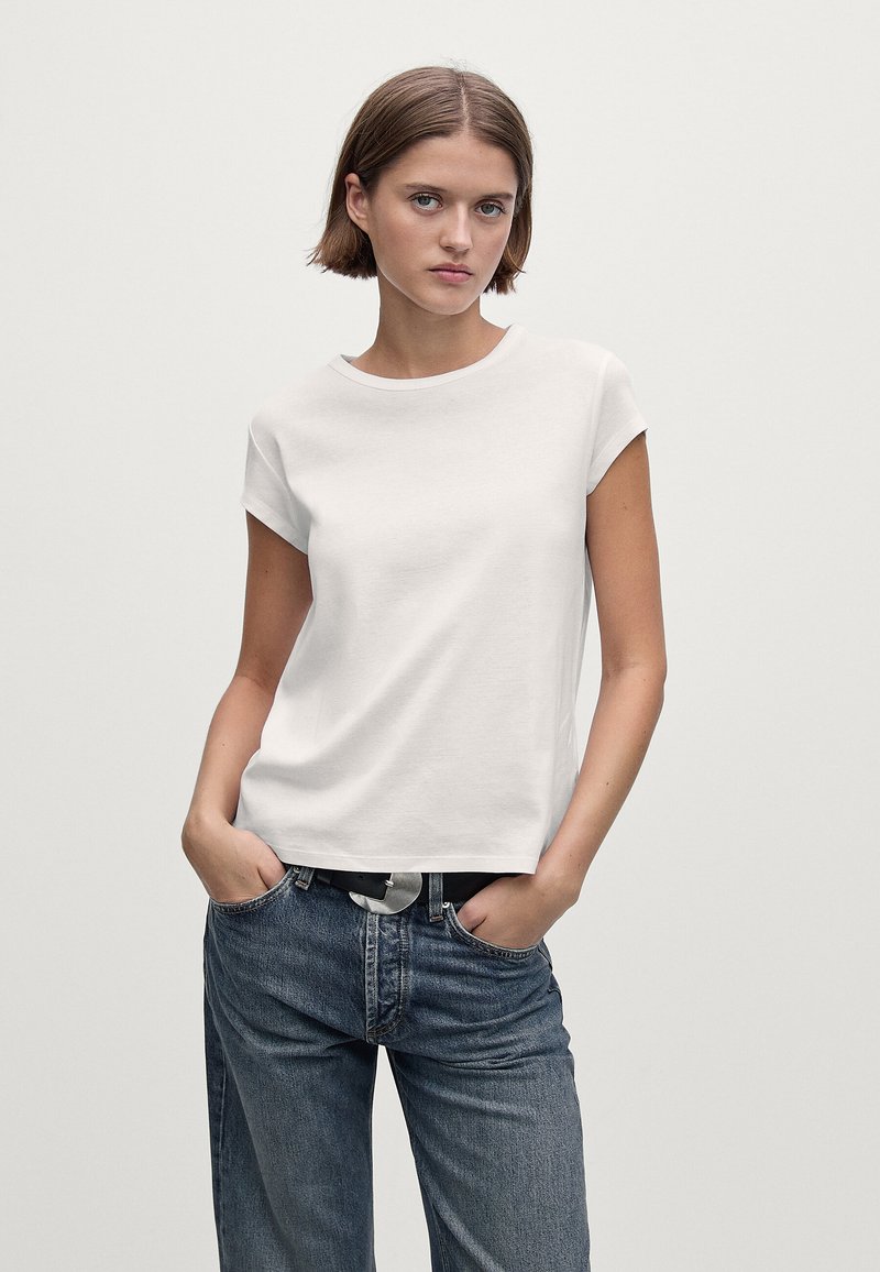 Donna con i capelli castani corti che indossa una maglietta bianca a maniche corte infilata in un paio di jeans blu larghi con una cintura nera, mani in tasca.