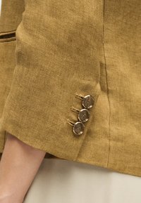 Εγγύς πλάνο του μανικιού ενός μουσταρδί λινό σακακιού με τρία μεταλλικά κουμπιά και ορατή ραφή κοντά σε ανοιχτόχρωμο δέρμα μπράτσο.