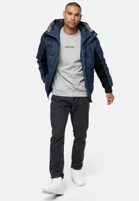 Marineblauwe pufferjack met zwarte accenten, ritszakken en een trekkoordkap, gedragen over een grijs sweatshirt en zwarte jeans, gecombineerd met witte sneakers.
